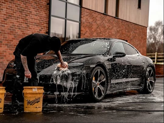 A person hand-washing a black car covered in foam